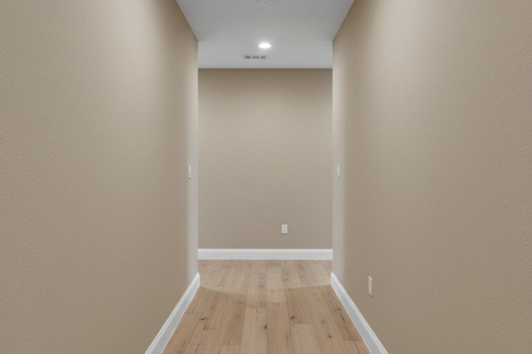 A brightly lit and empty L-shaped hallway in a modern home with neutral beige walls, white baseboards, and light oak laminate flooring. The space is evenly illuminated by recessed ceiling lights.