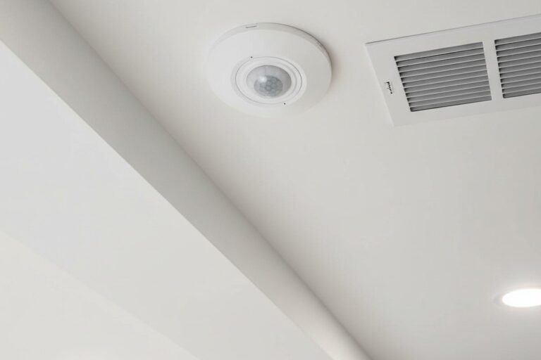 A low-angle shot looking up at a small, round, white motion sensor flush-mounted on a drywall ceiling bulkhead, positioned next to a square metal air register.