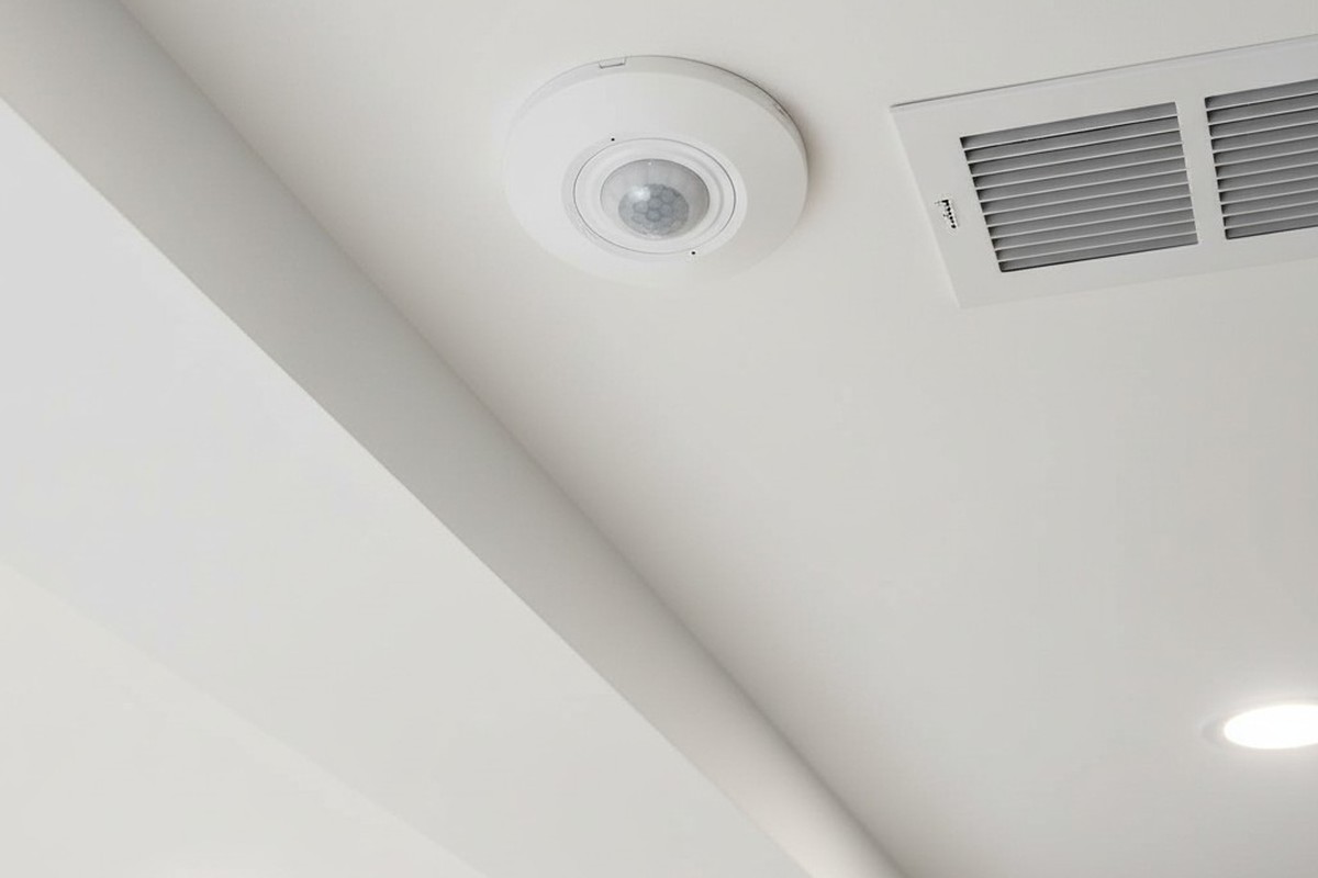 A low-angle shot looking up at a small, round, white motion sensor flush-mounted on a drywall ceiling bulkhead, positioned next to a square metal air register.