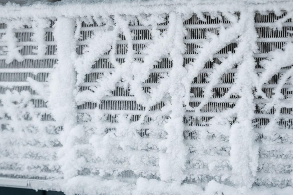 A close-up of an air conditioner's evaporator coil completely encased in a thick, jagged layer of white ice and frost, obscuring the metal fins.
