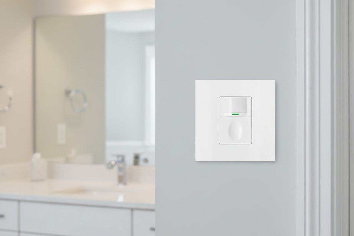 A white vacancy sensor switch is neatly installed on a light gray wall next to a door frame in a clean, modern bathroom with a vanity blurred in the background.