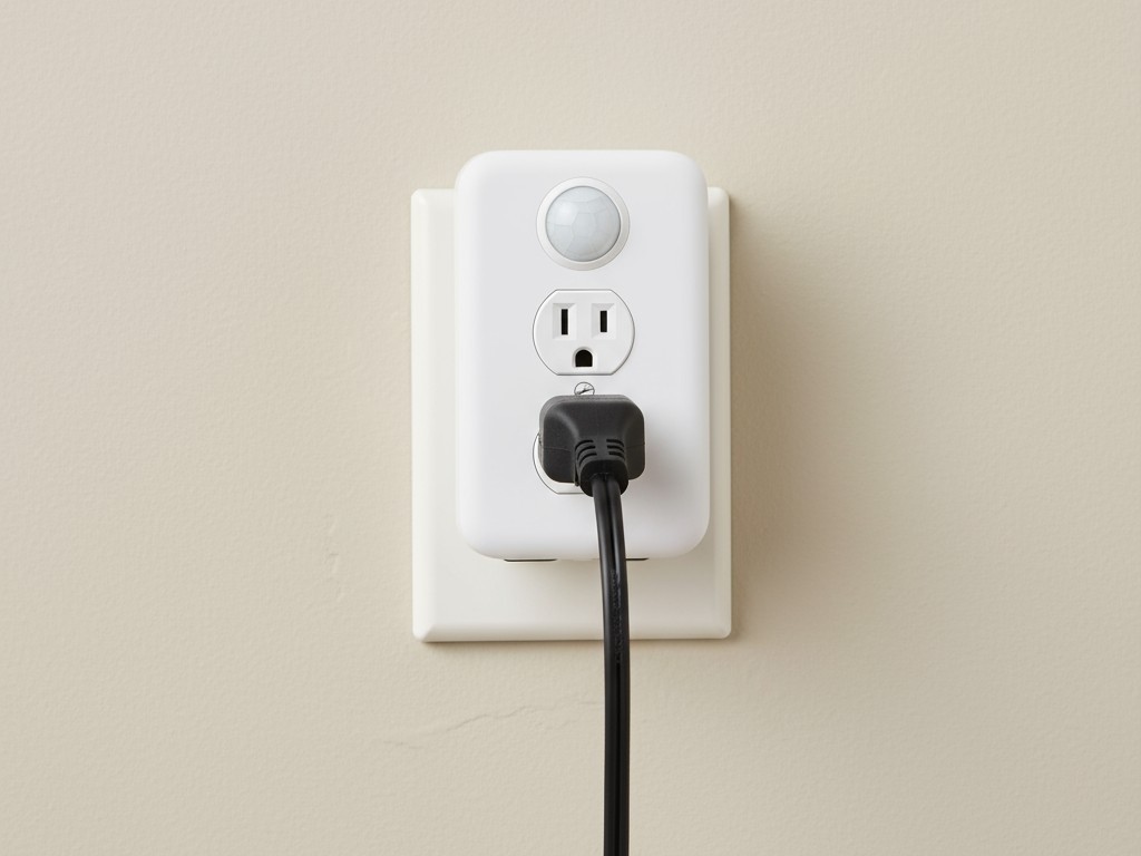 Un enchufe inteligente con sensor de movimiento conectado a una toma de corriente, con un cable de alimentación de un calentador insertado, mostrando una configuración de automatización sencilla.