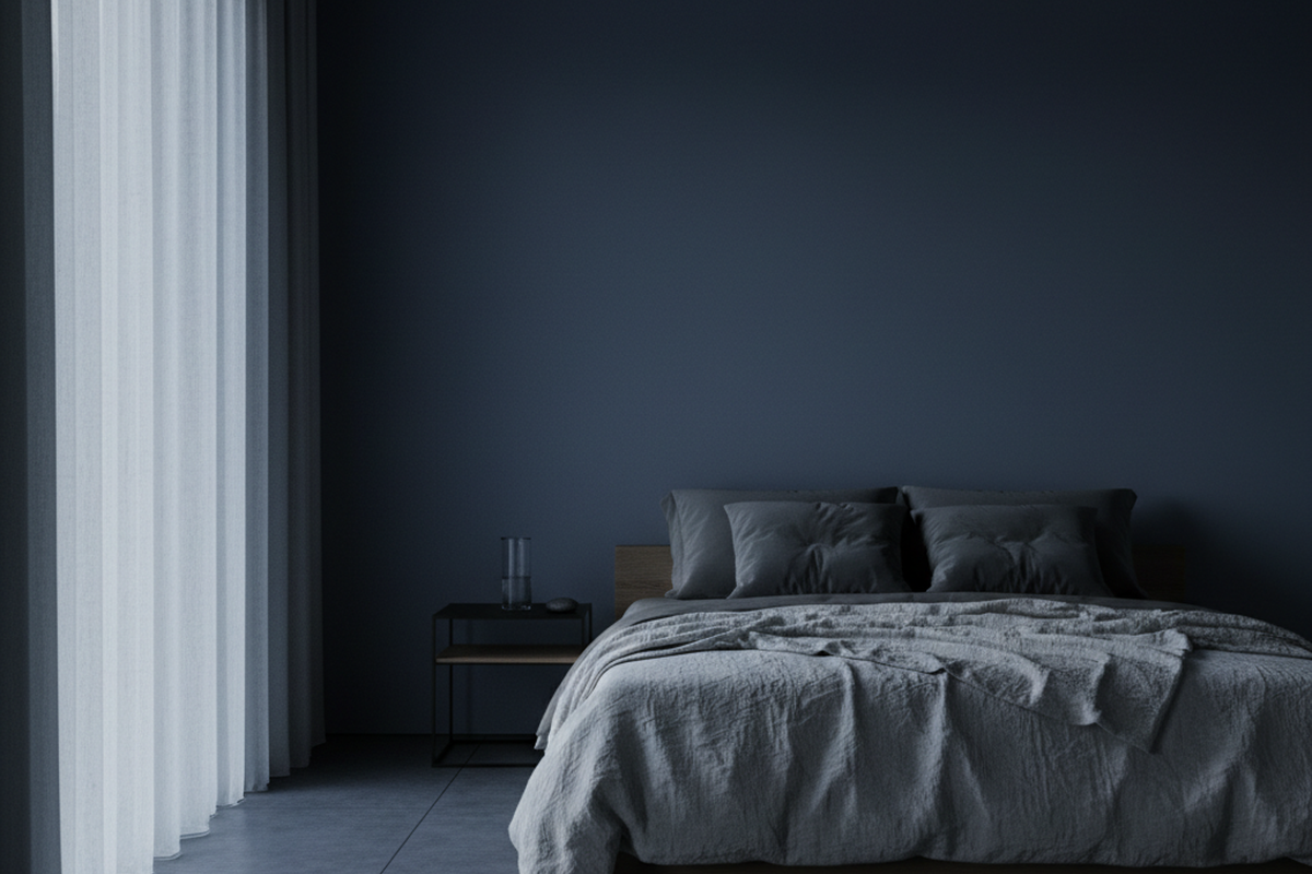A dimly lit bedroom features a low bed with grey linens and a glass of water on a side table against dark blue walls. Soft light filters through sheer floor-to-ceiling curtains, illuminating the room without artificial fixtures.