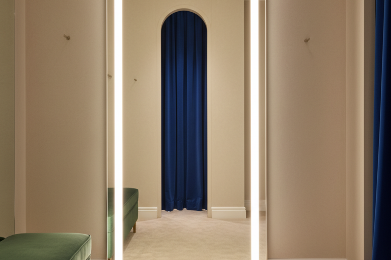 큰 전신 거울 양쪽 가장자리에 밝은 수직 LED 조명 스트립이 달려 있으며, 베이지색 방에 녹색 벨벳 벤치가 놓여 있다. 반사된 모습에는 짙은 파란색 커튼으로 덮인 아치형 출입구가 보인다.