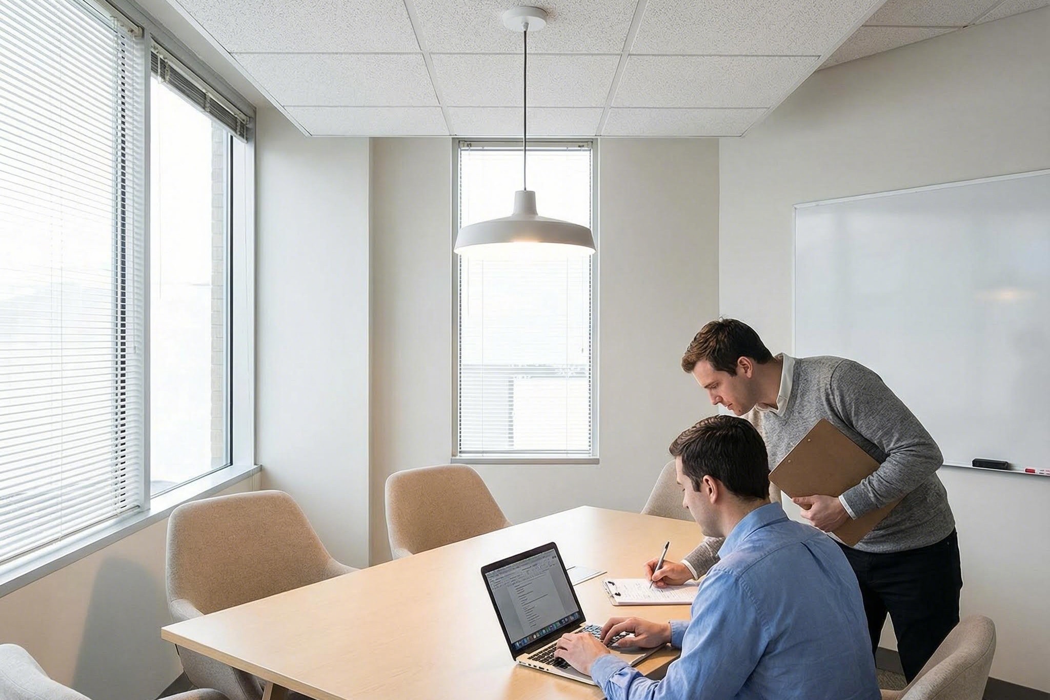 A small conference room table with laptops and a person seated while another person marks a simple grid map on a clipboard beneath a ceiling sensor.